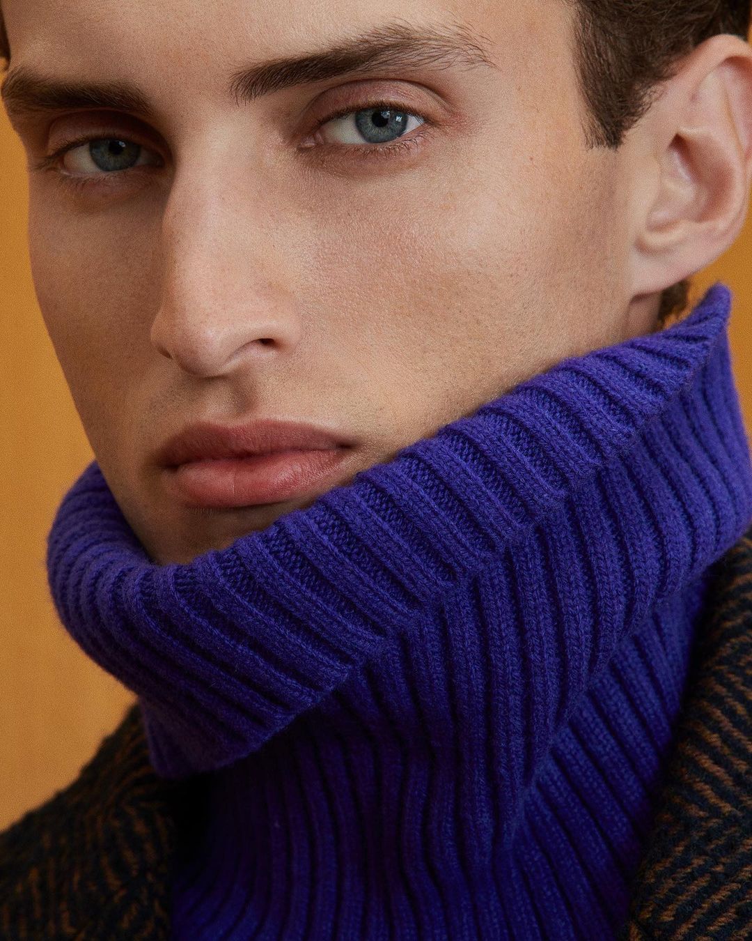 Close-up portrait of a fair-skinned adult male with blue eyes wearing a purple turtleneck sweater and a brown herringbone coat.