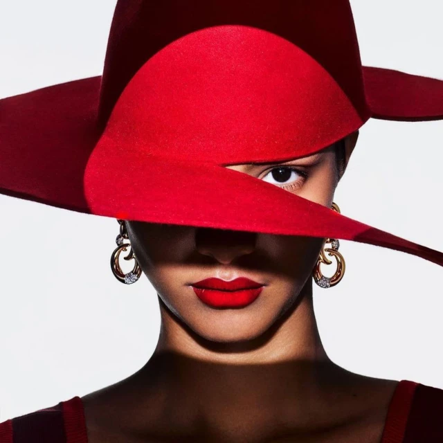 Close-up of a stylish adult female wearing a large red hat, gold earrings, and red lipstick against a bright white background.