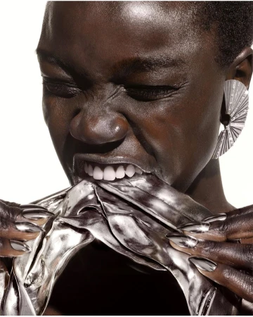 Close-up portrait of a Black woman biting a reflective metallic fabric with silver claw-like fingernails and a large fan earring