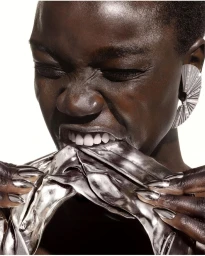 Close-up portrait of a Black woman biting a reflective metallic fabric with silver claw-like fingernails and a large fan earring