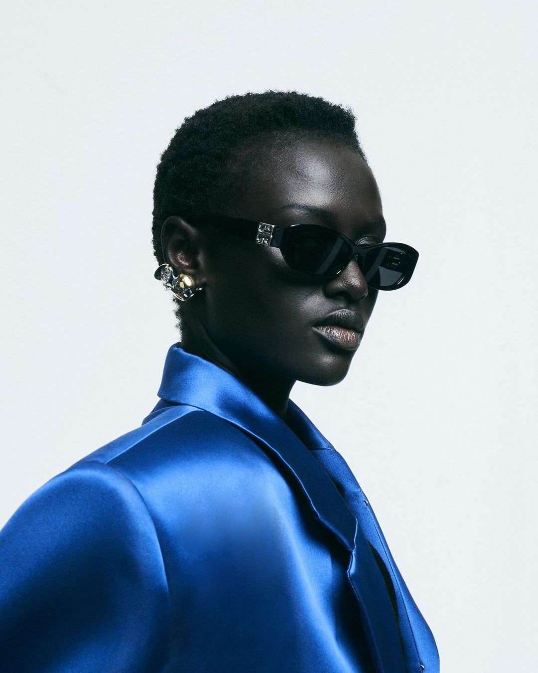 Fashion shot of a dark-skinned woman with short hair wearing black sunglasses and a shiny blue jacket against a white backdrop.