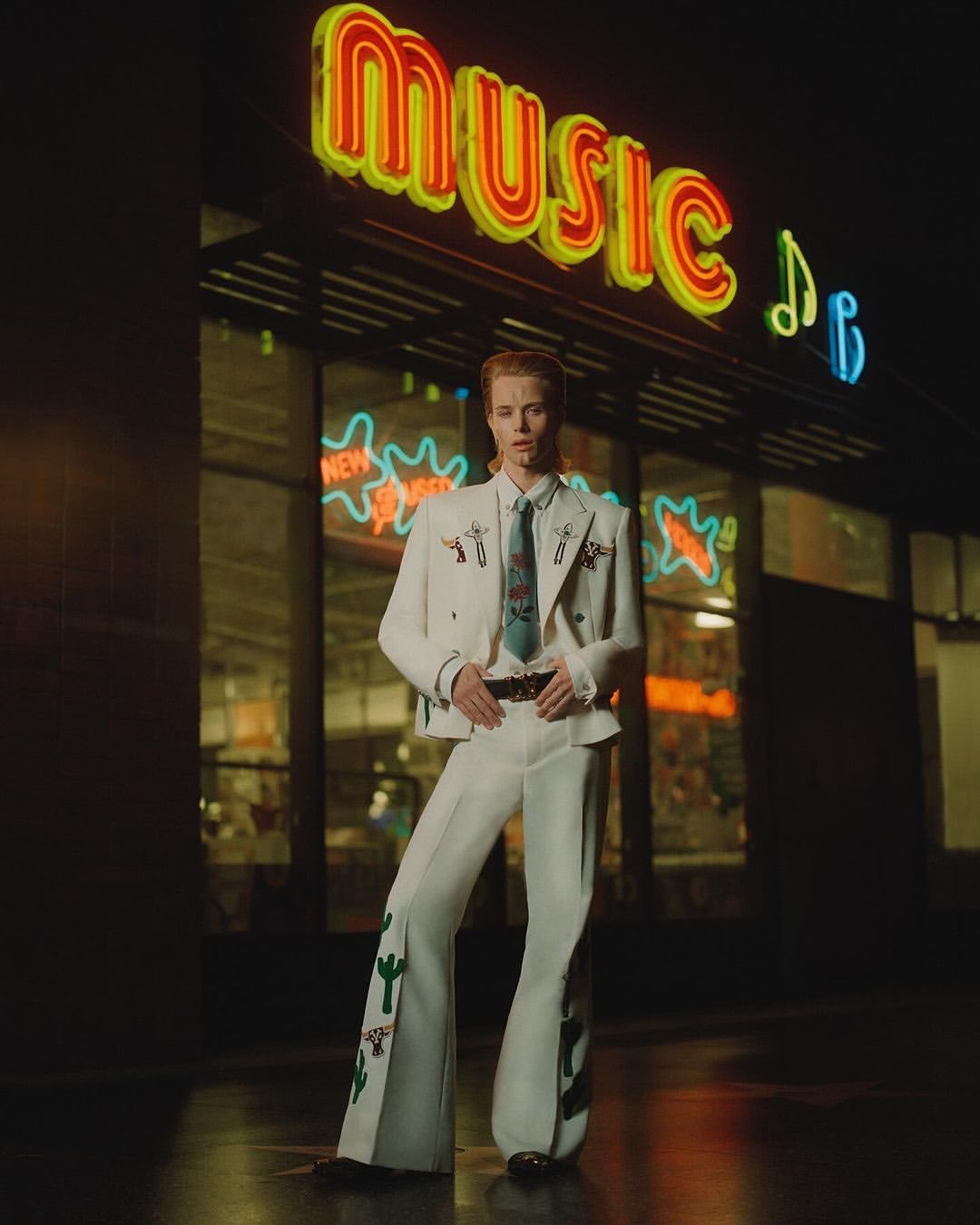 Man in white western suit poses in front of a neon-lit music shop at night, showcasing fashion and entertainment vibe.