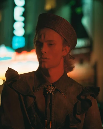 Close-up portrait of a stylish adult male in a vintage ruffled shirt and hat against an urban neon-lit cityscape backdrop at night.