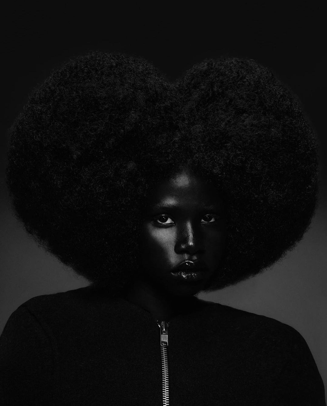 Dramatic black-and-white portrait of a woman with an afro hairstyle wearing a zip-up jacket, emphasizing dark tones and textures.