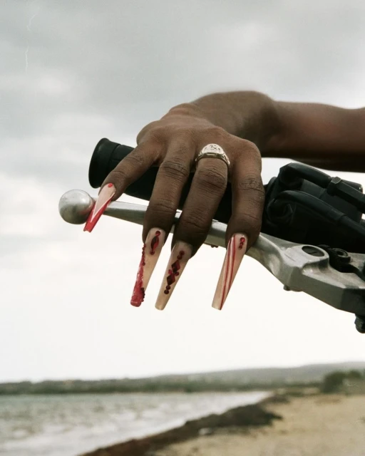Close-up of an adult's hand on a motorcycle handlebar displaying an intricate ring and long decorative nails with red accents outdoors.