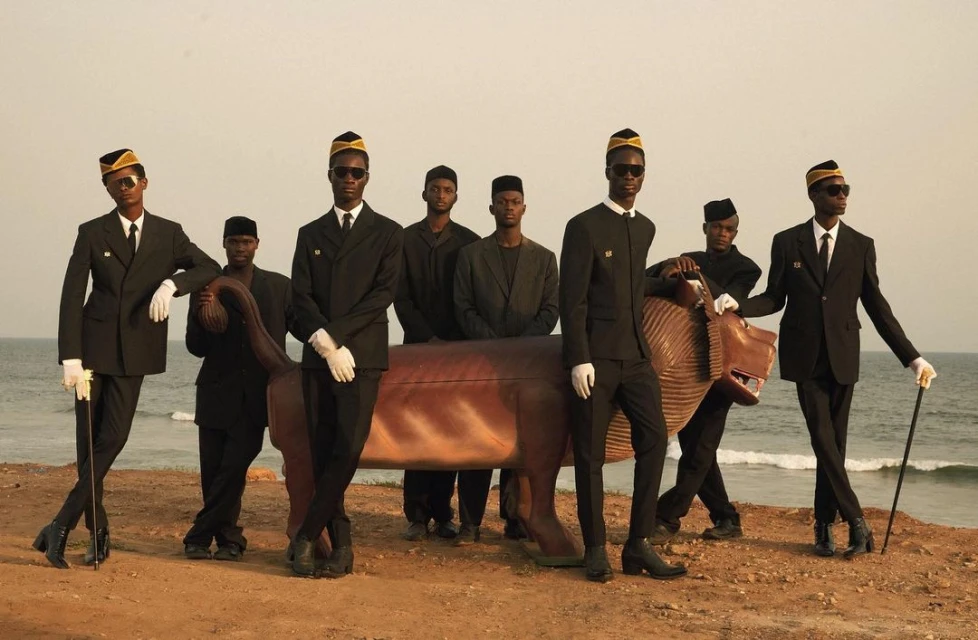 Group of seven black men in black suits and hats pose near a lion statue by the sea on a sandy shore, conveying a formal and stylish scene.