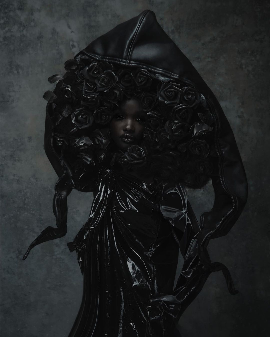 Studio portrait of a Black woman wearing a dark high-fashion hooded gown adorned with rose appliques against a textured grey backdrop.