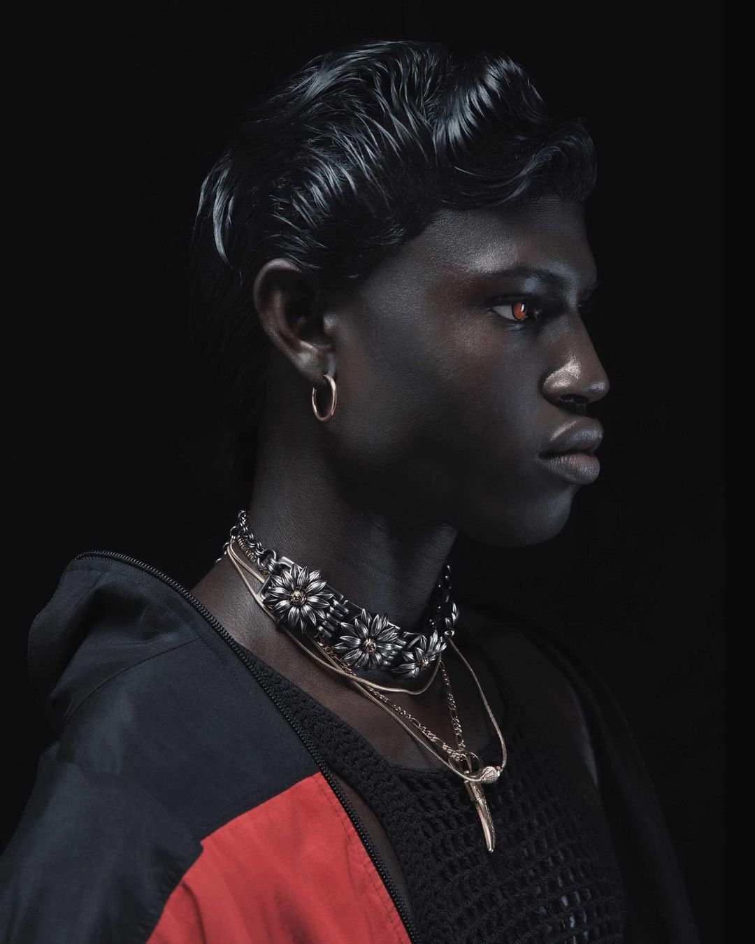 Portrait of a dark-skinned person with slicked-back wavy hair, silver hoop earring, and multiple layered necklaces, on a black background.