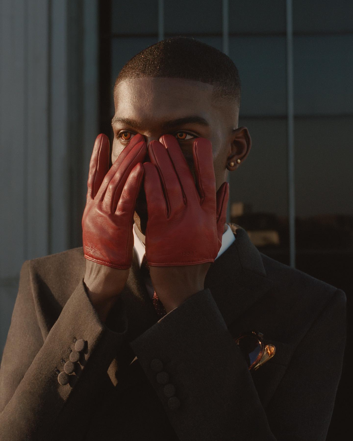Portrait of an adult Black male with short hair, wearing a dark suit and red leather gloves, holding his hands to his face.