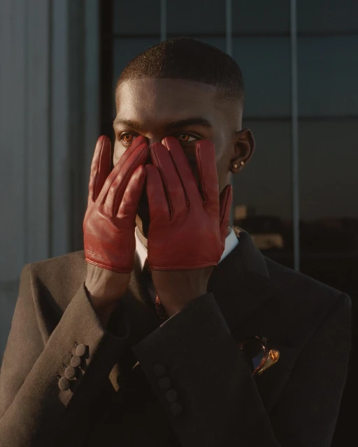 Portrait of an adult Black male with short hair, wearing a dark suit and red leather gloves, holding his hands to his face.