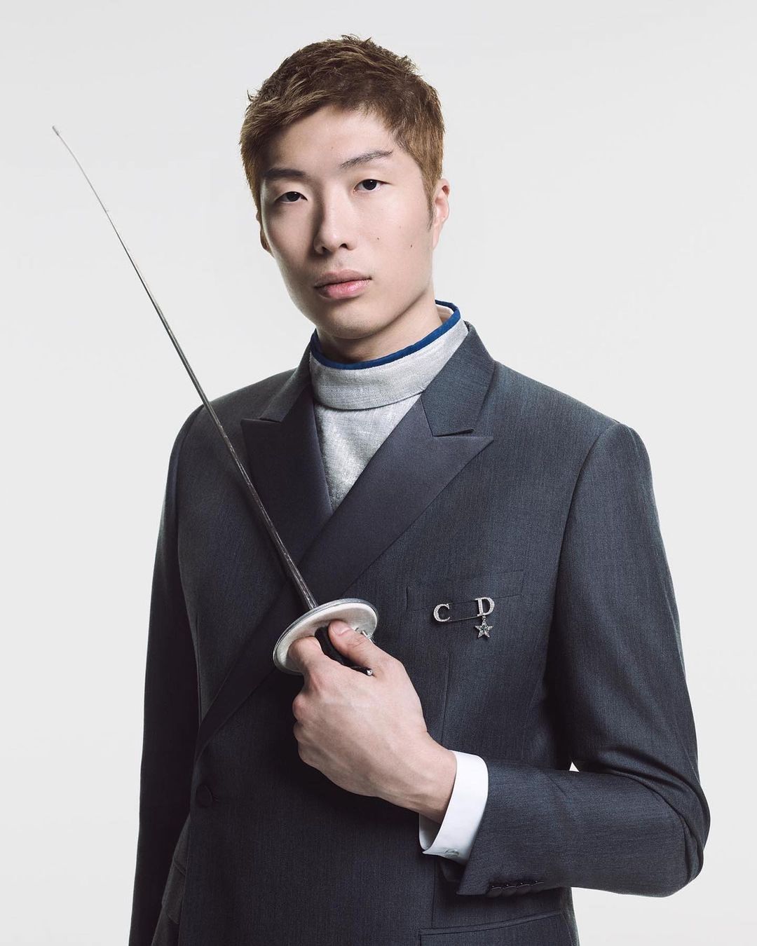 Portrait of a young Asian male model in a dark suit, holding a fencing foil against a white background with soft, even lighting.