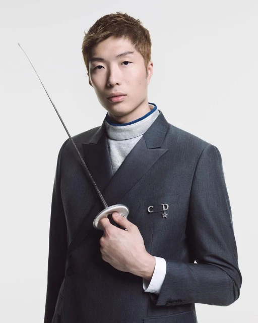 Portrait of a young Asian male model in a dark suit, holding a fencing foil against a white background with soft, even lighting.