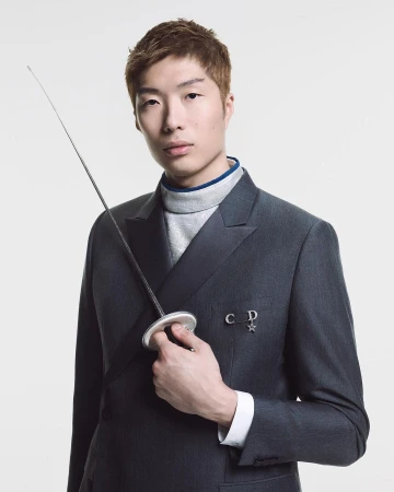 Portrait of a young Asian male model in a dark suit, holding a fencing foil against a white background with soft, even lighting.