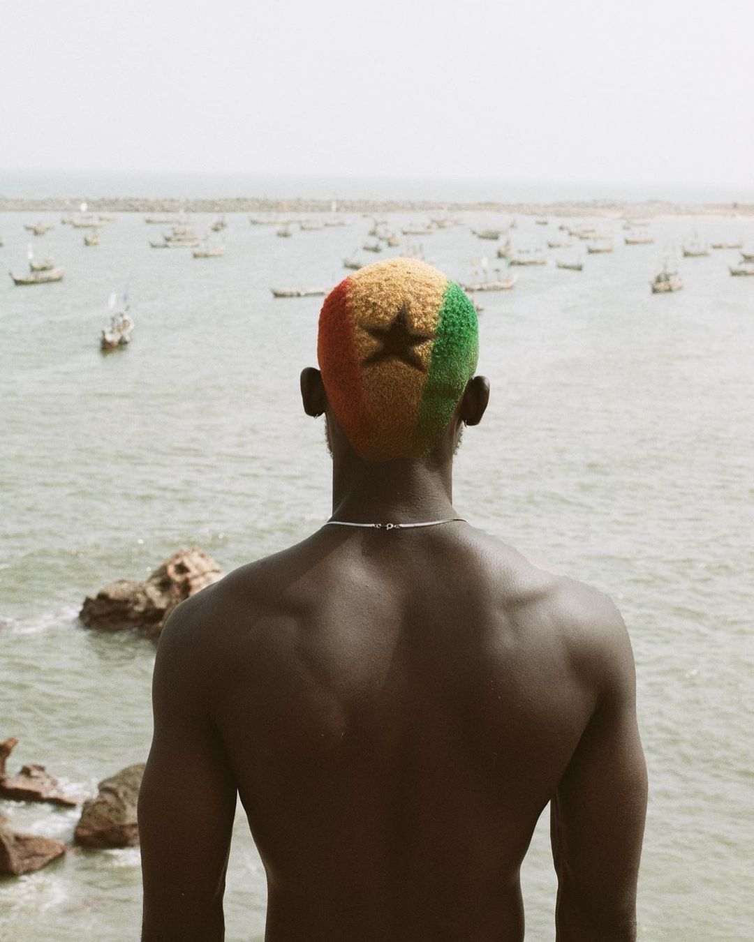 Back view of a shirtless adult male with a haircut dyed with the colors of the Ghanaian flag, staring at the sea and distant boats.