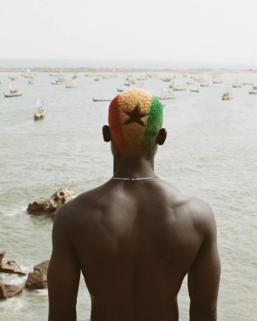 Back view of a shirtless adult male with a haircut dyed with the colors of the Ghanaian flag, staring at the sea and distant boats.