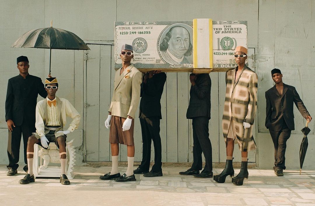 Stylish group of six Black men outdoors posing with a large prop dollar bill and black umbrellas in fashion editorial photography.