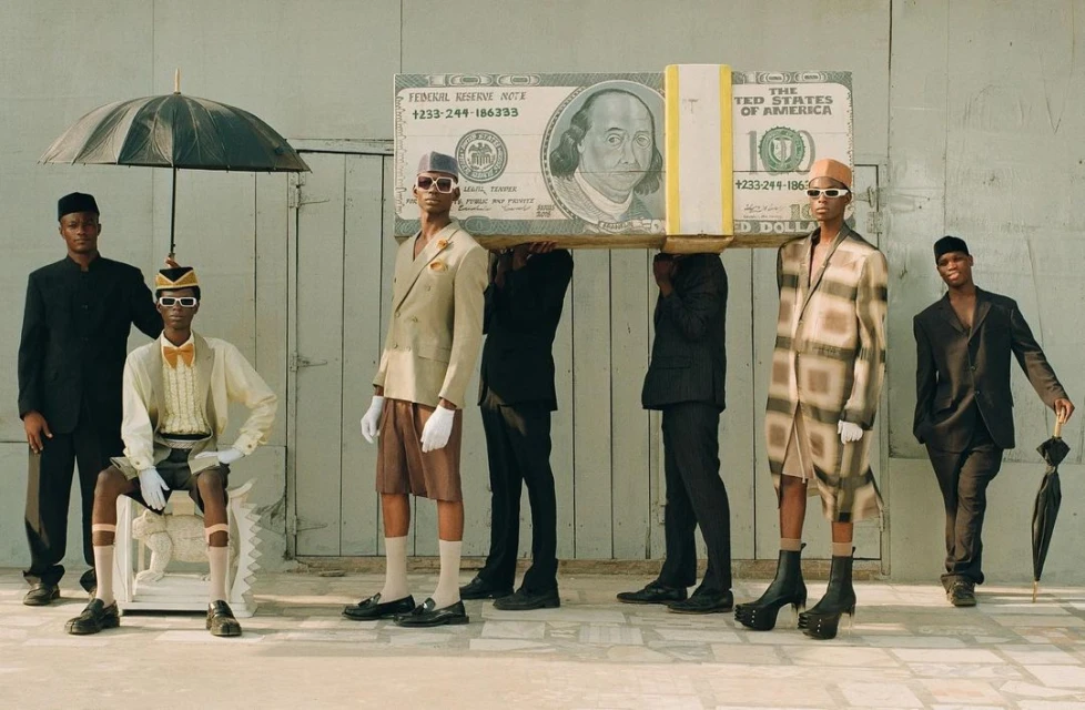 Stylish group of six Black men outdoors posing with a large prop dollar bill and black umbrellas in fashion editorial photography.
