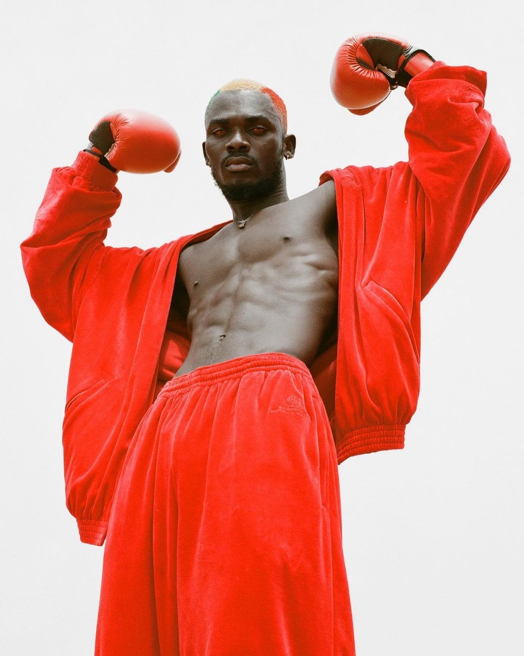 Athletic dark-skinned male boxer with orange hair stands with fists raised in red boxing gloves and a matching red velvet outfit.