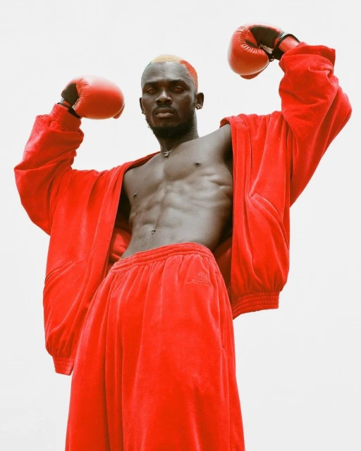 Athletic dark-skinned male boxer with orange hair stands with fists raised in red boxing gloves and a matching red velvet outfit.