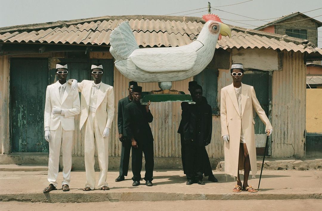 A group of people are carrying a large white chicken statue in front of a corrugated metal building with a weathered, textured exterior.