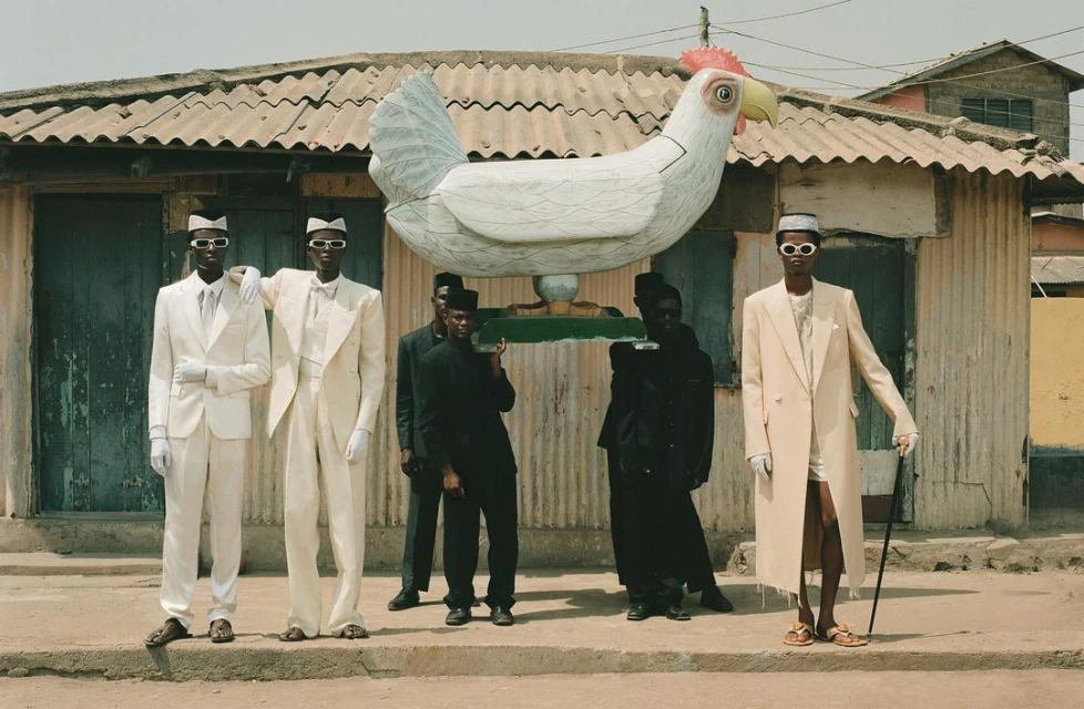 A group of people are carrying a large white chicken statue in front of a corrugated metal building with a weathered, textured exterior.