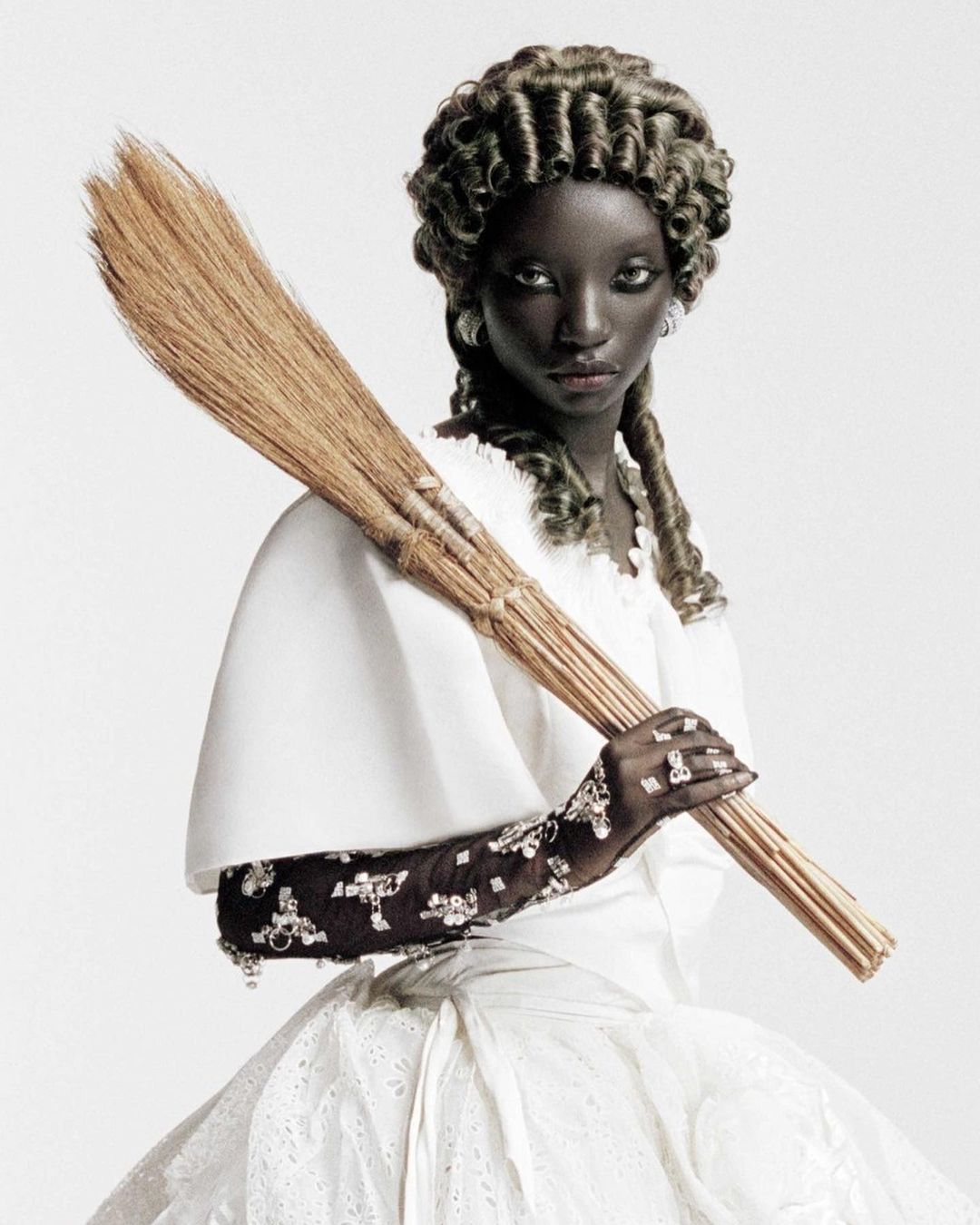 Dark-skinned woman with broom in white dress and formal curls. Fashion editorial style photo with unusual pose against white background.