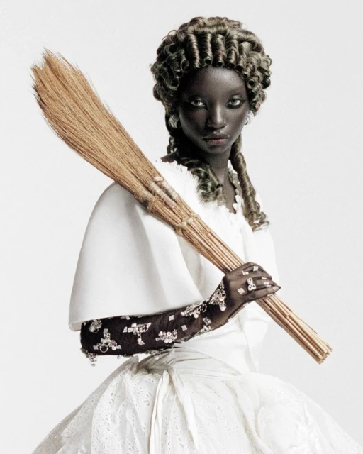 Dark-skinned woman with broom in white dress and formal curls. Fashion editorial style photo with unusual pose against white background.