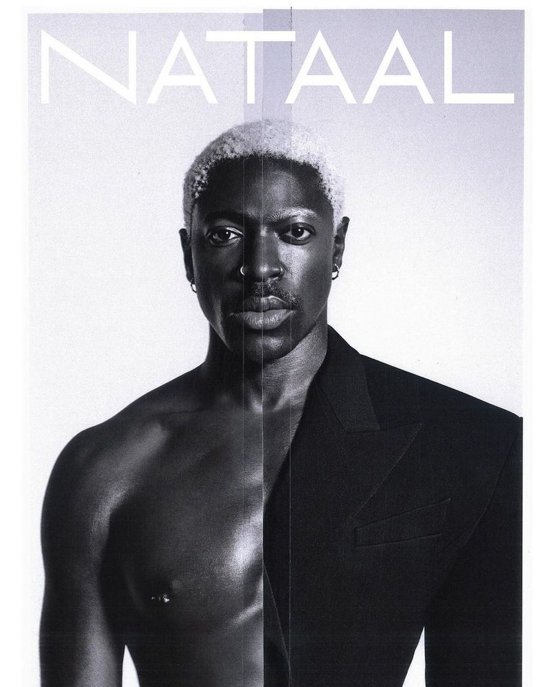 Striking black and white portrait of a shirtless Black man with platinum blonde hair wearing a blazer, set against a clean, light background.