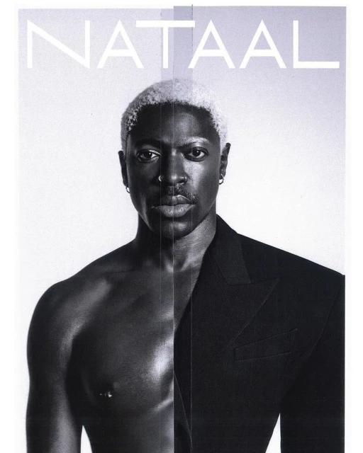 Striking black and white portrait of a shirtless Black man with platinum blonde hair wearing a blazer, set against a clean, light background.