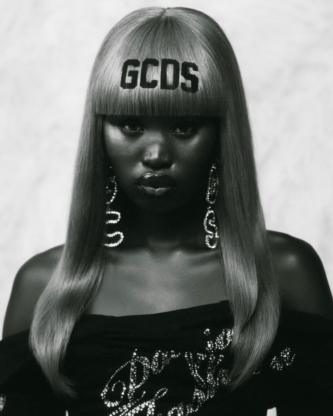 Portrait of Black model with blond hair and bangs styled with a GCDS logo, wearing an off-shoulder top and large earrings in a studio shot.
