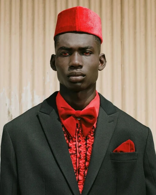 Portrait of a Black man in a black suit, red bowtie and fez-style hat, facing forward against a neutral wall, fashionably styled and lit.