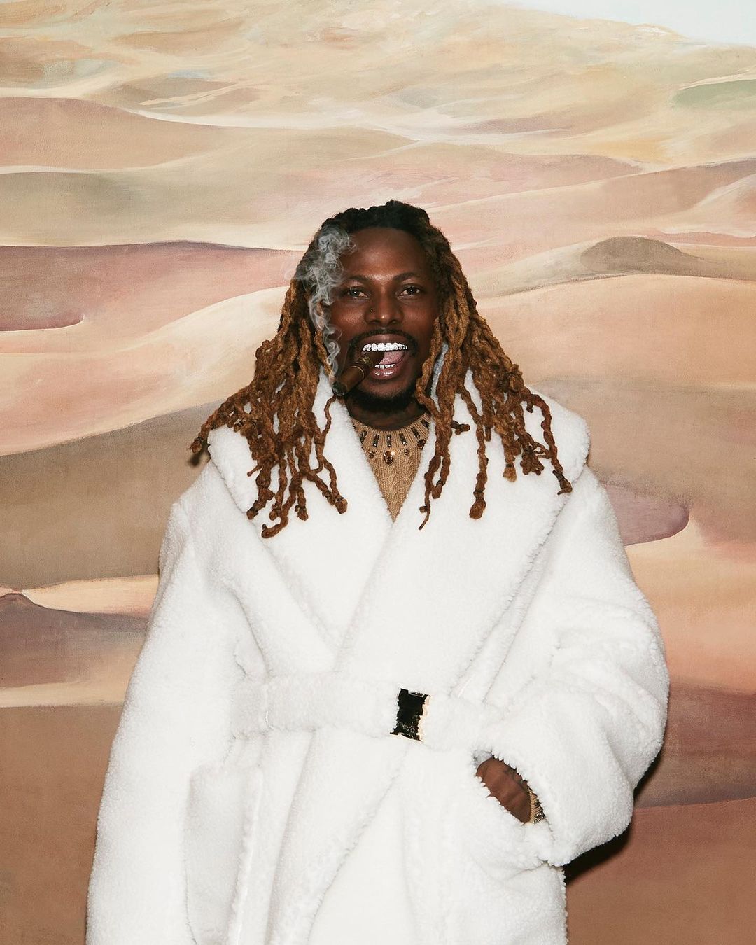 Smiling man with dreadlocks, wearing a white robe and holding a cigar, posing in front of a desert landscape painting.
