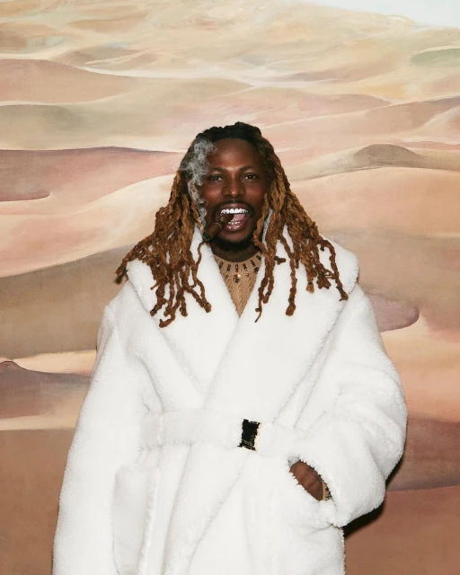 Smiling man with dreadlocks, wearing a white robe and holding a cigar, posing in front of a desert landscape painting.