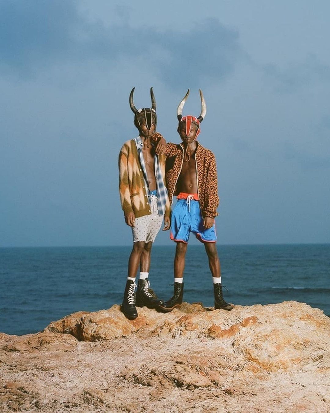 Two men stand on rocks wearing shorts, jackets, and horned masks against a sea and sky background, showcasing a fashion editorial.