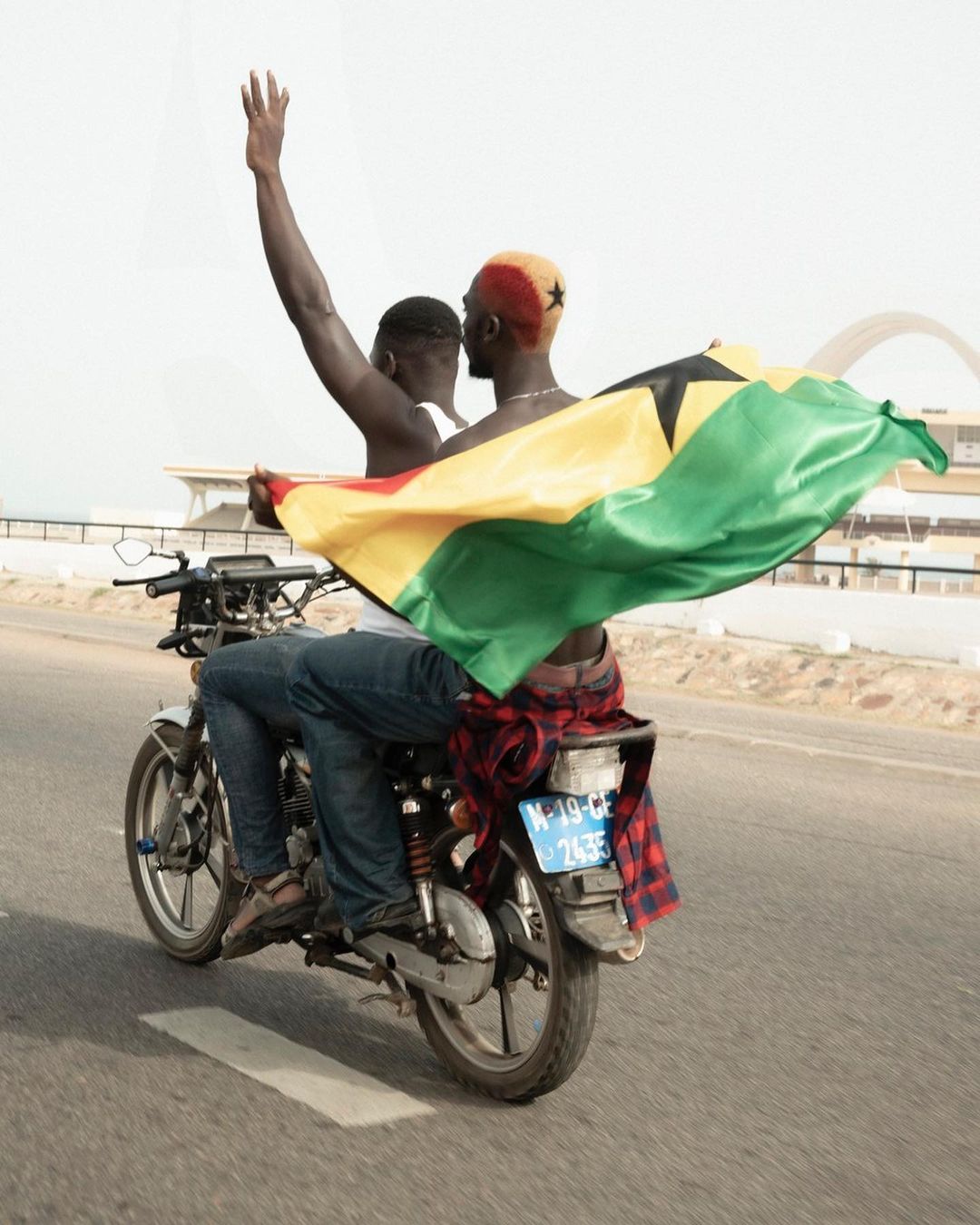 Two African men ride a motorcycle on a road with the Ghana flag blowing in the wind. One man raises his arm in celebration.
