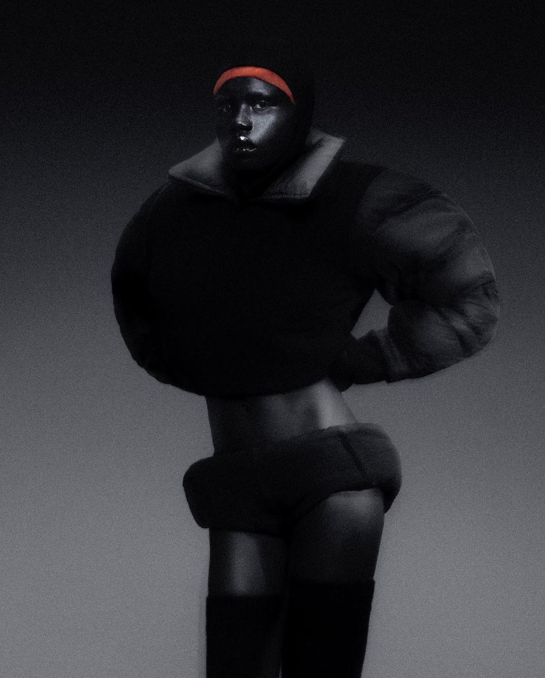 Dark-skinned person in a dark outfit and head covering poses in a studio, showing skin at midriff and upper thigh.