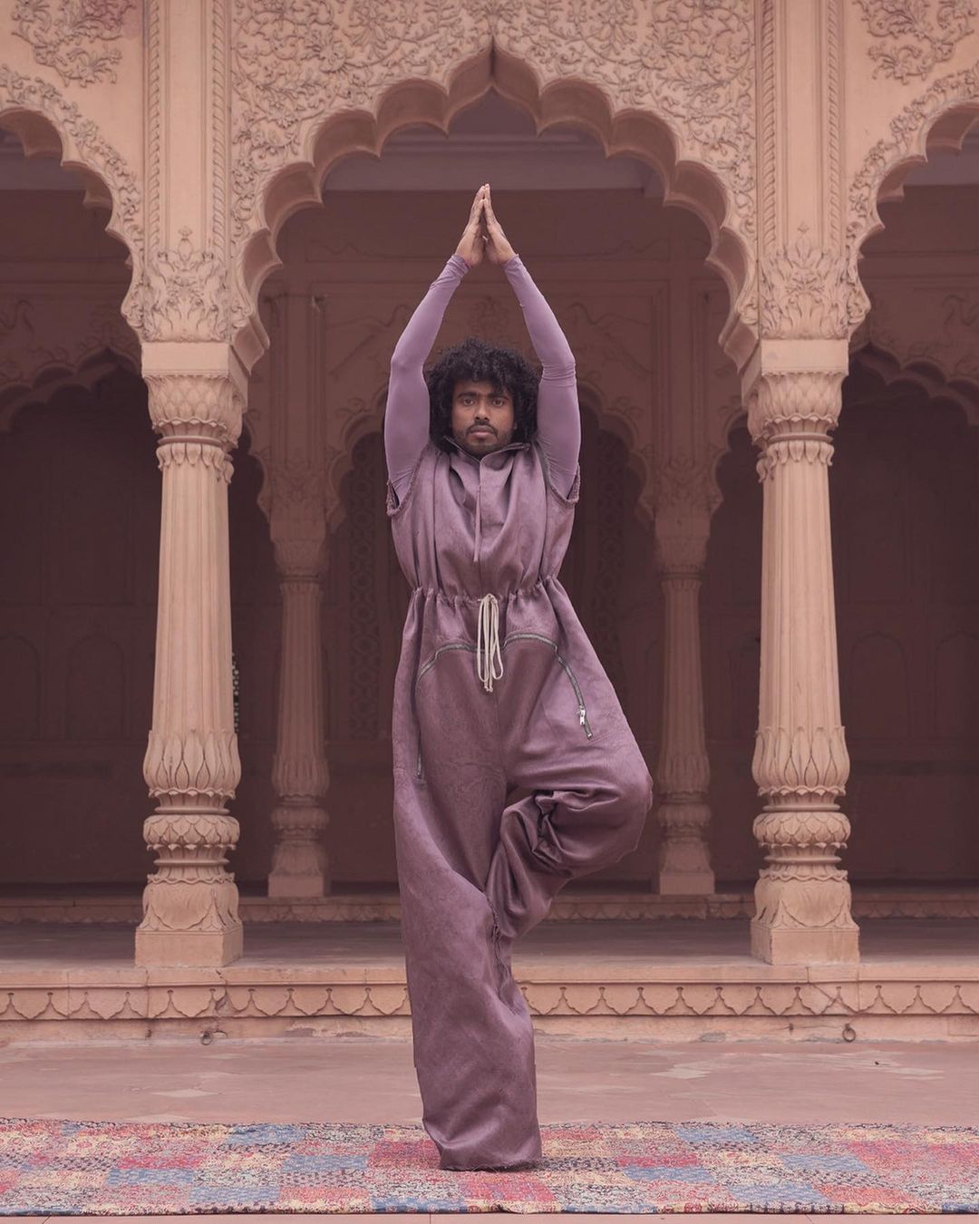 Man in a purple jumpsuit doing a yoga tree pose outdoors in front of ornate architecture, with hands raised above his head.