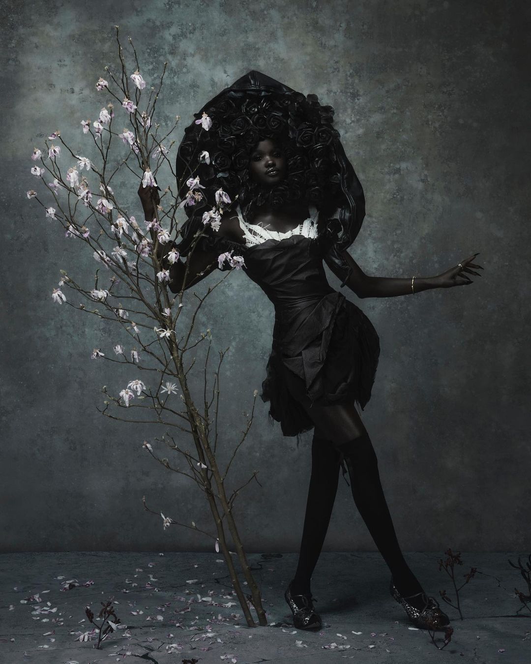 An African American woman is posing in a dark dress and sculptural headpiece, standing next to a small blossoming tree in a moody, grey studio.