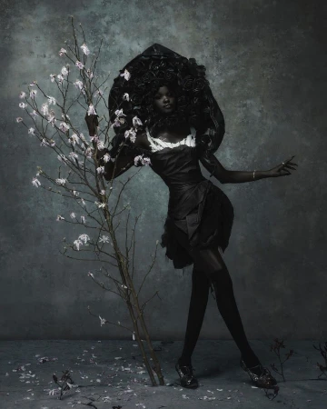 An African American woman is posing in a dark dress and sculptural headpiece, standing next to a small blossoming tree in a moody, grey studio.