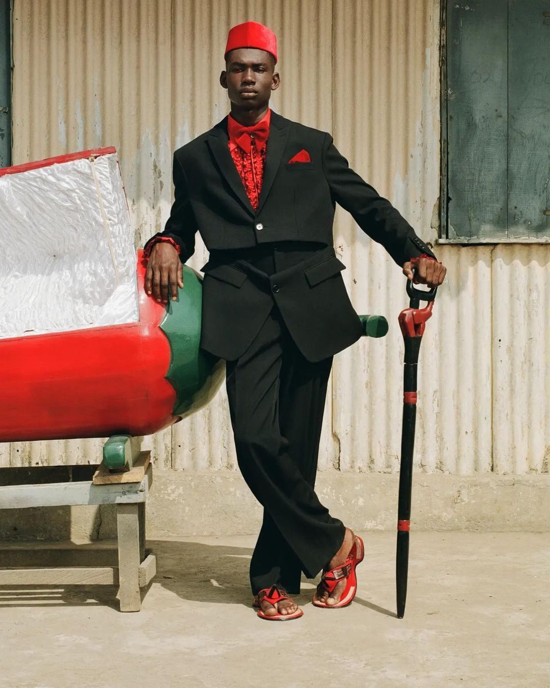 A stylish adult male poses outdoors wearing a black suit, red shirt, red hat, and red sandals next to a bench with a large red prop.