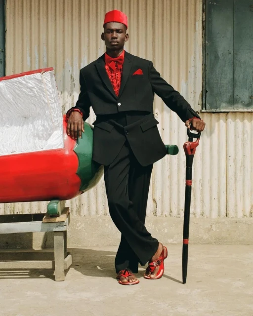 A stylish adult male poses outdoors wearing a black suit, red shirt, red hat, and red sandals next to a bench with a large red prop.