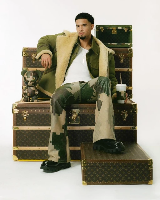 Man in stylish camo pants and jacket, posing on a Louis Vuitton trunk stack with a collectible toy and drink in a white studio.