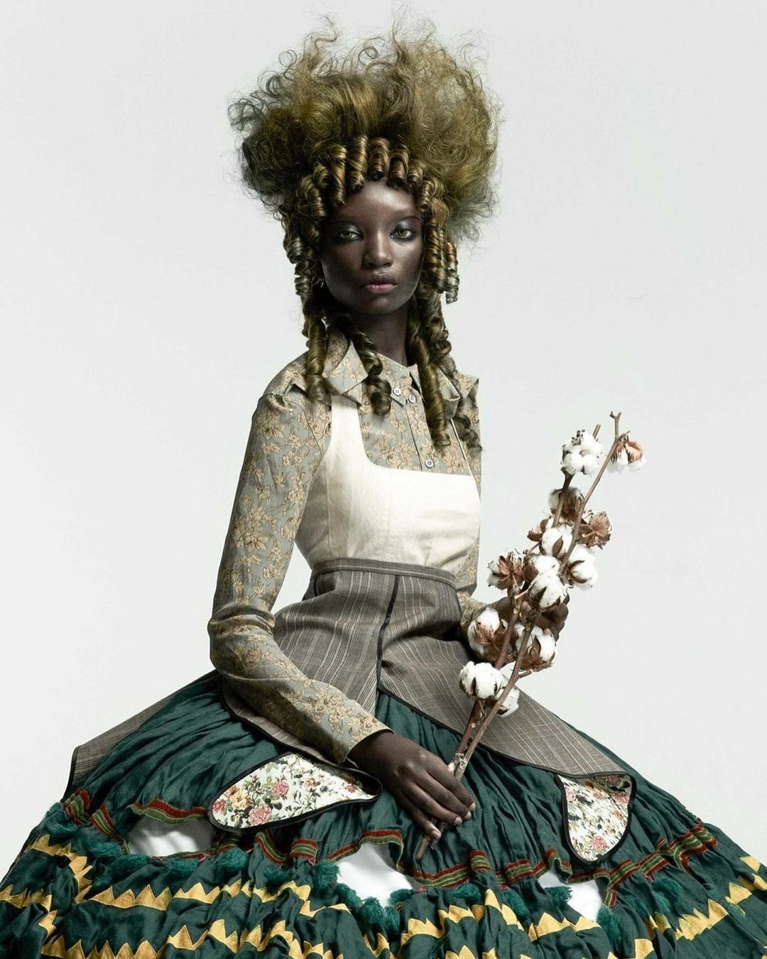 A dark-skinned woman wearing a voluminous green dress, elaborate curly hairstyle, and period costume is holding cotton branches in a studio shot.