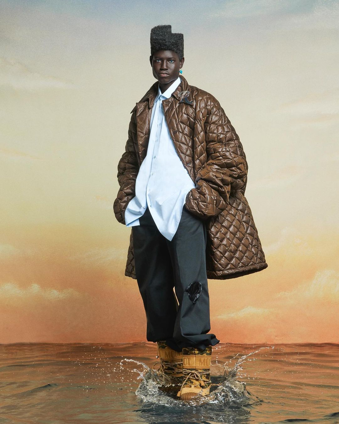 A dark-skinned model stands in ankle-deep water wearing a brown quilted coat, light blue shirt, dark pants, and winter boots, with a water background.