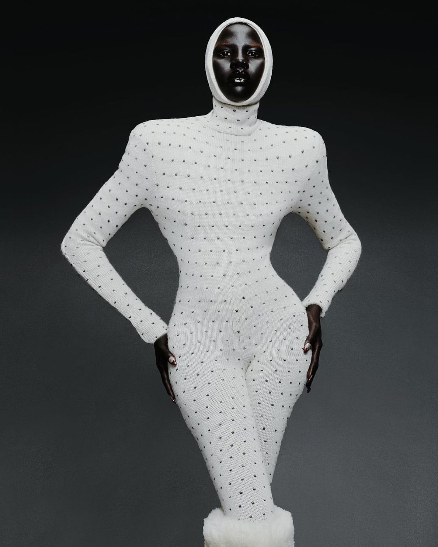 A woman with dark skin poses in a full-body, long-sleeved, dotted, white bodysuit and matching head covering against a grey background.