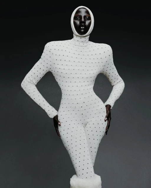 A woman with dark skin poses in a full-body, long-sleeved, dotted, white bodysuit and matching head covering against a grey background.