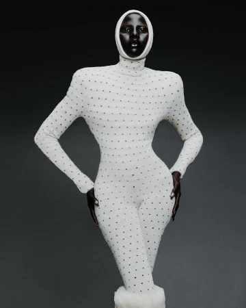 A woman with dark skin poses in a full-body, long-sleeved, dotted, white bodysuit and matching head covering against a grey background.