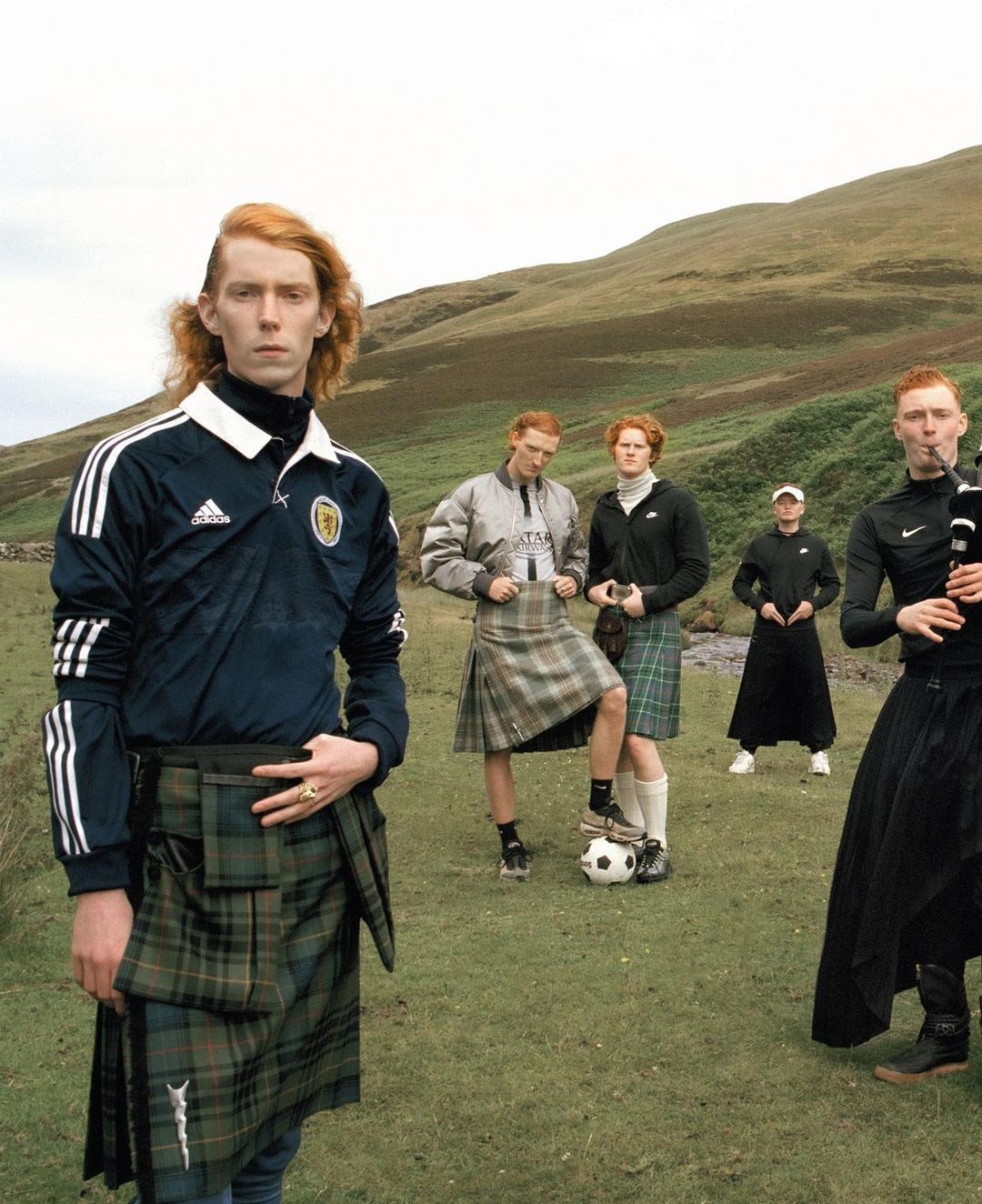 Group of red-haired male models wearing soccer jerseys and kilts pose in a grassy Scottish field with rolling hills in the background.