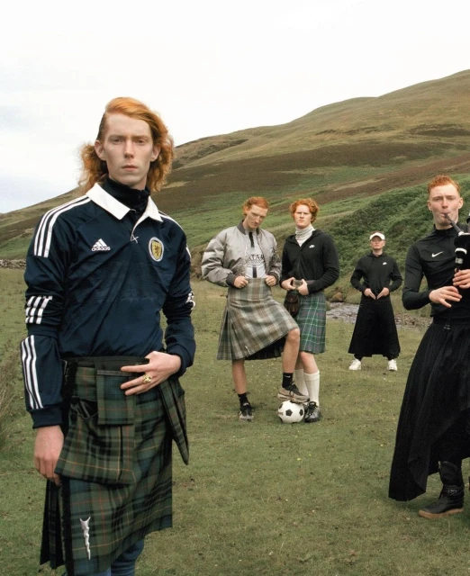 Group of red-haired male models wearing soccer jerseys and kilts pose in a grassy Scottish field with rolling hills in the background.