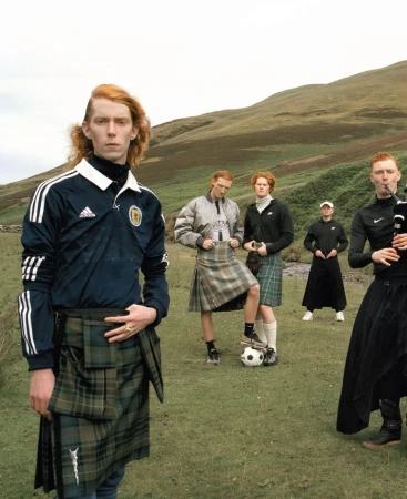Group of red-haired male models wearing soccer jerseys and kilts pose in a grassy Scottish field with rolling hills in the background.